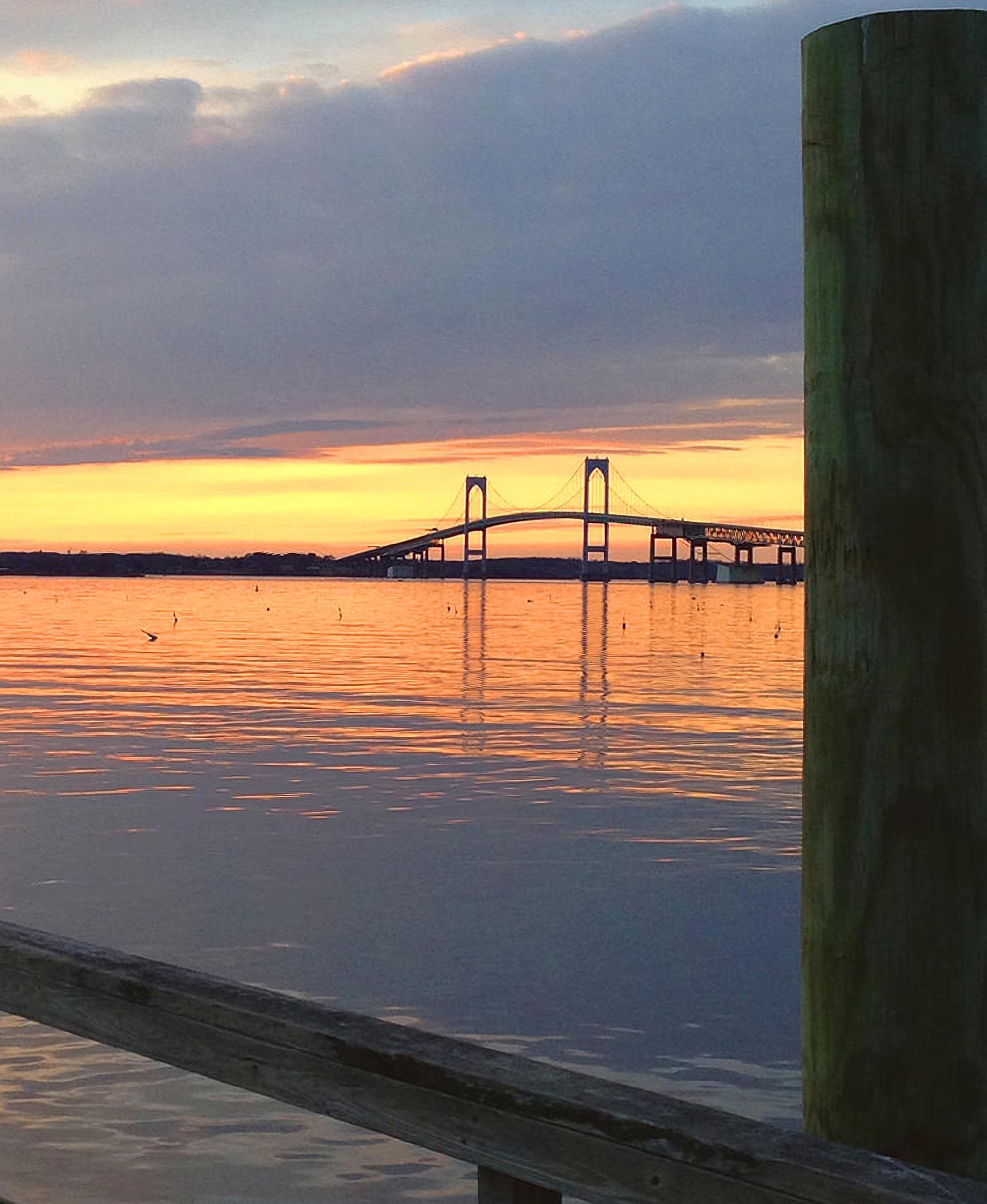A suspension bridge over the water at sunset, seen past a dock piling